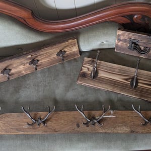 May include: A collection of rustic wooden coat racks with metal hooks. The racks display different designs, including antler-shaped hooks, traditional hooks, and decorative handles. The wood has a natural, distressed finish, giving a vintage feel.