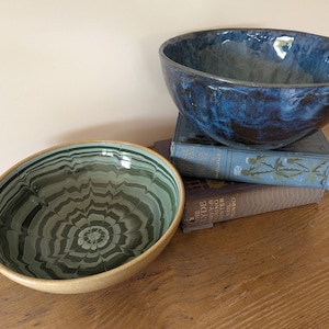 May include: Two ceramic bowls. One bowl features a deep blue glaze with dark mottled tones and a smooth, rounded rim. The second bowl displays a light green interior with a dark green, radiating, marbled pattern and a tan, unglazed exterior rim.