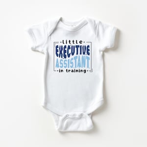 Peut inclure: Un body pour bébé blanc à manches courtes avec un motif graphique portant l'inscription "little EXECUTIVE ASSISTANT in training" en noir et dans des tons de bleu. Le texte est encadré par un fin rectangle noir avec de petites étoiles.