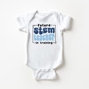 May include: A white short-sleeved baby bodysuit featuring a graphic design with the text "future STEM teacher in training" in dark blue and light blue wavy lettering, enclosed within a black rectangular frame with small star accents.
