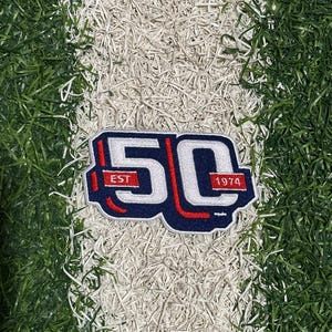 May include: A navy blue and white patch featuring the number "50" in a bold font, with red outlining. The patch includes the text "EST 1974" and is set against a backdrop of green and white artificial turf.