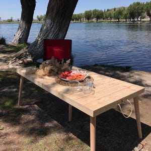 May include: A light-colored wooden picnic table with a red portable record player, fruit, and glasses set up near a lake. The table has a rope handle and is supported by four angled legs.