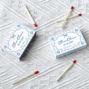 May include: Two matchboxes with blue and white striped patterns and seashell illustrations. The text "Ethan + Olivia 05.20.2027 Miami, FL" is printed on the boxes. Several matches with red tips are scattered around the boxes.