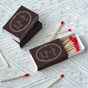 May include: Two dark brown matchboxes with white decorative oval frames and the text Amelia & Henry, 09.12.2027, SC, Charleston. One box is closed, and the other is open, revealing wooden matches with red tips. Loose matches with red tips are scattered.