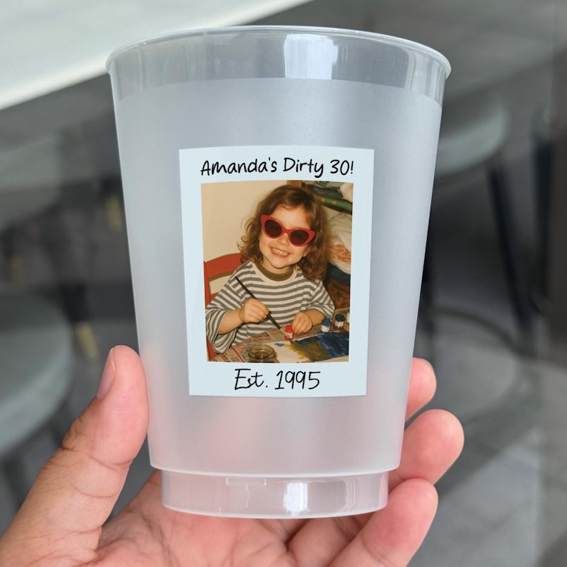 K&ouml;nnte beinhalten: Ein gefrosteter Plastikbecher mit einem Fotoetikett. Das Etikett zeigt ein Kinderfoto einer Person mit Sonnenbrille, mit dem Text "Amanda's Dirty 30!" und "Est. 1995". Der Becher wird von einer Hand gehalten.