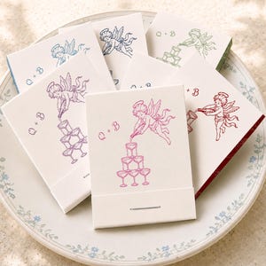 May include: A collection of rectangular matchboxes with white covers, each featuring a line art illustration of a winged cherub pouring a bottle into a champagne tower. The text Q + B is printed in various colors including pink, blue, and green.