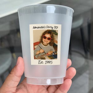 K&ouml;nnte beinhalten: Ein durchscheinender Plastikbecher mit einem Foto einer jungen Person mit Sonnenbrille. Das Foto tr&auml;gt die Aufschrift "Amanda's Dirty 30!" und "Est. 1995". Der Becher wird von einer Hand gehalten.