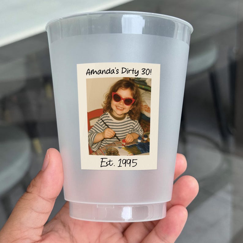 K&ouml;nnte beinhalten: Ein durchscheinender Plastikbecher mit einem Foto einer jungen Person mit Sonnenbrille. Das Foto tr&auml;gt die Aufschrift "Amanda's Dirty 30!" und "Est. 1995". Der Becher wird von einer Hand gehalten.