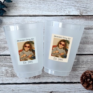 K&ouml;nnte beinhalten: Zwei gefrostete Plastikbecher mit einem Foto einer Person mit Sonnenbrille und dem Text "Amanda's Dirty 30!" und "Est. 1995". Die Becher stehen auf einer Holzoberfl&auml;che.