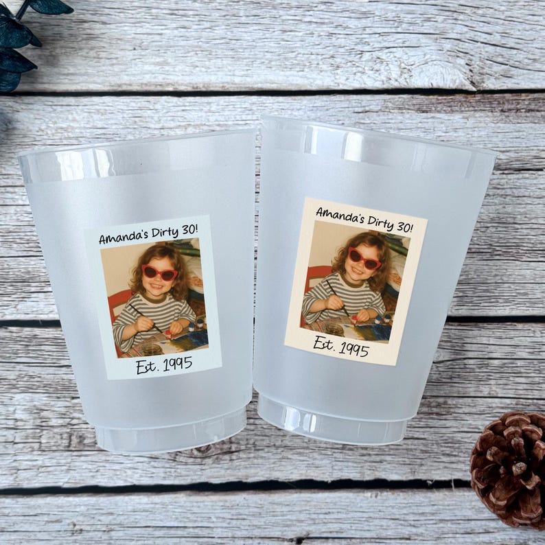 K&ouml;nnte beinhalten: Zwei gefrostete Plastikbecher mit einem Foto einer Person mit Sonnenbrille und dem Text "Amanda's Dirty 30!" und "Est. 1995". Die Becher stehen auf einer Holzoberfl&auml;che.
