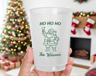 May include: A frosted plastic cup with a green illustration of Santa Claus holding a drink and a bottle. The words "HO HO HO" and "The Wilson's" are printed on the cup. The background includes a Christmas tree and a fireplace.