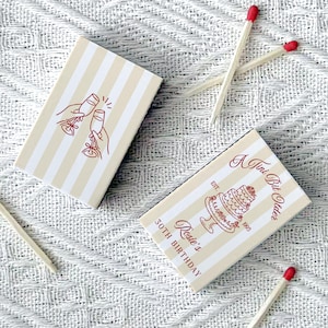 May include: Two matchboxes with cream and white stripes. One box has a red illustration of two hands toasting champagne glasses. The other box has a cake illustration and text that reads "A Tiny Bit Older, Rosie's 30th Birthday".