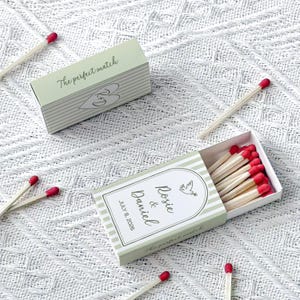 May include: Two matchboxes with the text "The perfect match" and a heart design. One box is open, revealing red-tipped matches. The other box is closed. The open box has the names "Rosie & Daniel" and the date "July 8, 2026".