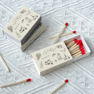 May include: Off-white matchboxes with black line art illustrations and the text "J + B" and "06/02/26". The matches have red tips. The text "The Perfect Match" is also on the boxes.