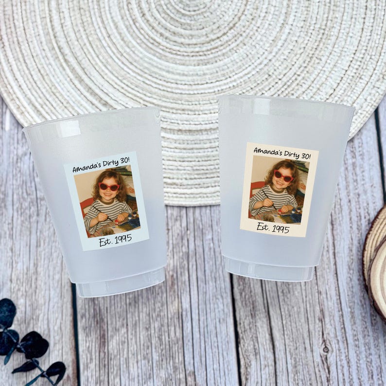 K&ouml;nnte beinhalten: Zwei gefrostete Plastikbecher mit einem Aufkleber mit einem Foto eines jungen M&auml;dchens mit Sonnenbrille. Der Aufkleber lautet "Amanda's Dirty 30!" und "Est. 1995". Die Becher stehen auf einer Holzoberfl&auml;che.