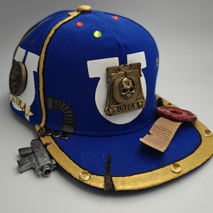 May include: A blue baseball cap featuring a white U-shaped emblem, a central bronze-coloured skull plaque with the text ULTRA, a small silver-coloured firearm replica, a scroll with a red wax seal, gold trim, and small colourful circular accents.
