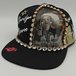 May include: Black baseball cap featuring a Marilyn Monroe image, embellished with rhinestones. The cap includes the text "Marilyn Monroe" and "Metropolitan Style The Big Apple". A red lip graphic is on the brim.
