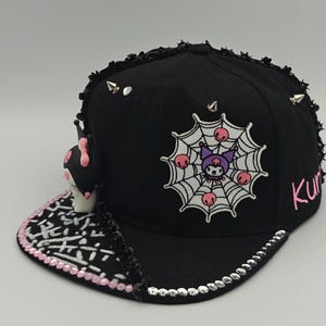 May include: Black baseball cap with a flat brim, featuring a spiderweb design with a cartoon character and the word "Kurumi" in pink. The cap is adorned with silver studs and a decorative trim.