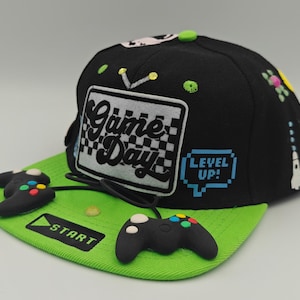 Puede incluir: Gorra de béisbol negra y verde con detalles de mando de videojuegos. La gorra presenta las palabras "Game Day" en la parte delantera, un gráfico "Level Up!" y una etiqueta "Start". La visera es de color verde brillante.