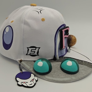 Könnte beinhalten: Eine weiße Baseballkappe mit einem violetten und rosafarbenen Buchstaben F, einem schwarzen Logo und zwei türkisfarbenen, kuppelförmigen Akzenten auf dem Schirm. Ein violett-weißer Charakteranhänger hängt an der Seite, der Schirm zeigt ein Muster.