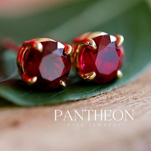 May include: A pair of round-cut, deep red gemstone stud earrings set in gold prongs. The jewelry is positioned on a green leaf. The text PANTHEON FINE JEWELRY is visible in white lettering at the bottom right of the image.