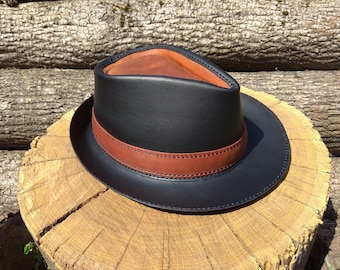 May include: A black leather fedora hat with a brown leather band and interior. The hat is sitting on a wooden surface, showcasing its classic design and craftsmanship. The hat's brim is approximately 2.5 inches wide.