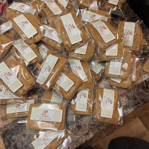Peut inclure: Plusieurs friandises rectangulaires emball&eacute;es individuellement, de couleur brun clair, possiblement du caramel ou du toffee, dispos&eacute;es sur une surface sombre. Chaque paquet porte une &eacute;tiquette avec du texte et un graphisme, probablement pour une baby shower.
