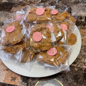 Peut inclure: Une assiette blanche pr&eacute;sente plusieurs morceaux de croquant aux noix de p&eacute;can emball&eacute;s individuellement. Chaque morceau est de couleur brun clair et scell&eacute; dans du plastique transparent. Des autocollants circulaires roses avec du texte en cursive sont appos&eacute;s sur l'emballage.