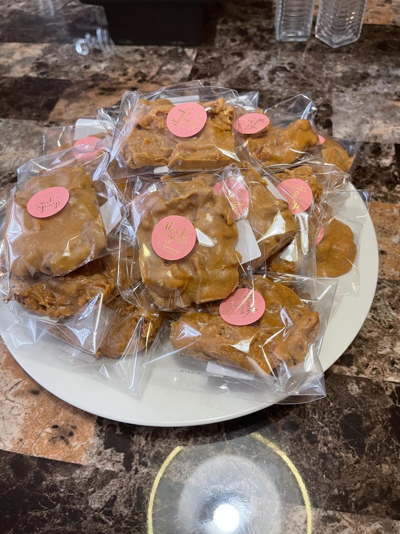 Peut inclure: Une assiette blanche pr&eacute;sente plusieurs morceaux de croquant aux noix de p&eacute;can emball&eacute;s individuellement. Chaque morceau est de couleur brun clair et scell&eacute; dans du plastique transparent. Des autocollants circulaires roses avec du texte en cursive sont appos&eacute;s sur l'emballage.