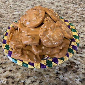 Peut inclure: Une assiette remplie de pralines aux noix de p&eacute;can. Les bonbons sont de couleur brun clair et ont un aspect lisse et brillant. L'assiette a une bordure color&eacute;e avec des formes de diamants violets, verts et dor&eacute;s.