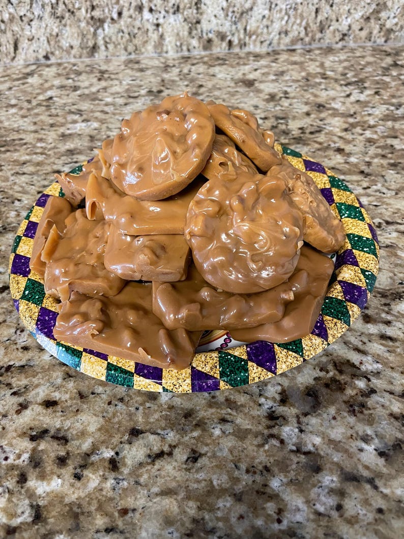 Peut inclure: Une assiette remplie de pralines aux noix de p&eacute;can. Les bonbons sont de couleur brun clair et ont un aspect lisse et brillant. L'assiette a une bordure color&eacute;e avec des formes de diamants violets, verts et dor&eacute;s.