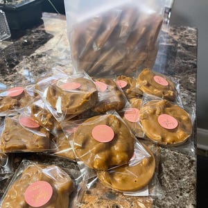 Peut inclure: Pralines aux noix de p&eacute;can emball&eacute;es individuellement, de couleur brun clair, avec des autocollants roses "Merci". Un grand sac de pralines similaires est en arri&egrave;re-plan. Les pralines sont dispos&eacute;es sur une surface &agrave; motifs de marbre.