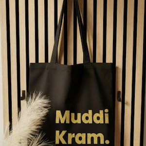 Op de afbeelding: Zwarte draagtas met gouden glittertekst "Muddi Kram". De tas hangt aan een zwarte haak tegen een houten lattenwand. Een decoratieve plant staat in de linker benedenhoek.