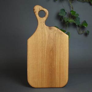 Può includere: Tagliere in legno marrone chiaro con manico con apertura circolare. La tavola ha una forma rettangolare con angoli arrotondati e un motivo a venature naturali del legno. Il manico è curvo, su sfondo grigio scuro.