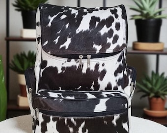 May include: A black and white cowhide backpack with multiple zipped compartments. The bag features a flap closure and adjustable straps. The backpack is made of a natural material and has a unique spotted pattern.