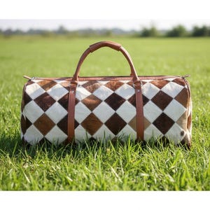 May include: A duffel bag featuring a geometric diamond pattern in shades of brown, white, and tan. It has brown leather handles and straps. The bag rests on green grass.