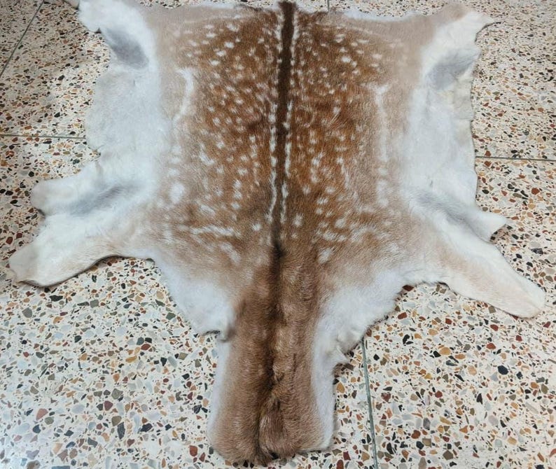May include: A natural deer hide rug, featuring a pattern of brown spots on a tan background. The hide has a central brown stripe and white edges, with the shape of the deer's body and legs visible.