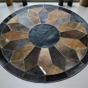 Könnte beinhalten: Ein runder Teppich mit geometrischem Design. Der Teppich hat einen zentralen dunkelblauen Kreis, umgeben von einem Muster aus braunen, grauen und beigen Formen. Die Formen sind mit hellfarbenem Faden zusammengenäht, und der äußere Rand ist dunkelgrau.