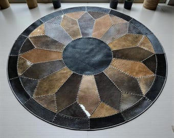 May include: A round rug with a geometric design. The rug features a central dark blue circle surrounded by a pattern of various brown, gray, and tan shapes. The shapes are stitched together with light-colored thread, and the outer edge is dark gray.