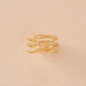 Op de afbeelding: Een set stapelbare ringen in goudkleur. De ringen hebben een textuur, organisch ontwerp. Eén ring is versierd met kleine, heldere edelstenen. De ringen worden gepresenteerd op een licht perzik-gekleurd oppervlak.