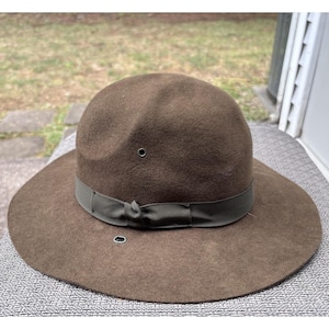 Könnte beinhalten: Brauner Filzhut mit breiter Krempe und dunkelgrünem Band. Der Hut hat zwei kleine Belüftungslöcher auf der Krone und eines am Rand. Ein klassisches Accessoire für verschiedene Anlässe.