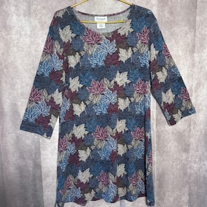 May include: A long-sleeve tunic top featuring a repeating pattern of maple leaves in shades of blue, purple, and grey on a dark heathered background. The garment has a round neckline and a Serenade brand label stitched at the inner collar.