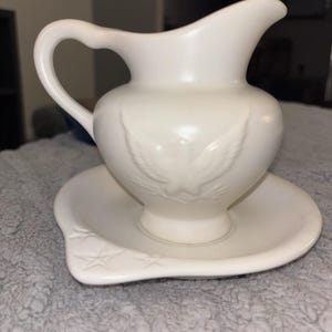 Peut inclure: Ensemble pichet et cuvette en céramique crème. Le pichet est doté d'une anse et d'un bec verseur, avec un motif d'aigle en relief sur le devant. Le bassin assorti présente un motif d'étoiles sur le côté.