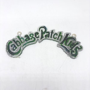 May include: A decorative sign featuring the text Cabbage Patch Kids in a stylised, arched font. The letters are filled with a glossy, dark green enamel finish and outlined in a metallic silver border, with two small circular loops for hanging.