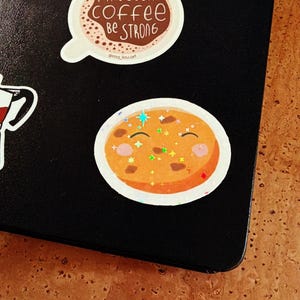 Könnte beinhalten: Zwei Aufkleber auf einer schwarzen Oberfläche. Ein Aufkleber ist eine Kaffeetasse mit dem Text "MAY YOUR COFFEE BE STRONG". Der andere Aufkleber ist ein lächelnder orangefarbener Kreis mit Glitzerakzenten.