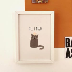 Könnte beinhalten: Weiß gerahmter Kunstdruck mit einer skurrilen Illustration einer schwarzen Katze mit einer Kaffeetasse auf dem Kopf. Der Text "ALL I NEED" steht über der Katze. Das Kunstwerk wird an einer weißen Wand mit anderen dekorativen Elementen ausgestellt.