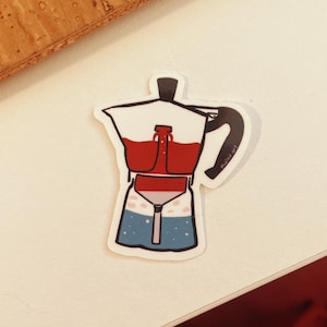Könnte beinhalten: Ein Aufkleber mit einer Cartoon-Illustration eines Espressokochers. Der obere Teil ist weiß mit einem schwarzen Griff, der mittlere Teil ist rot und der untere Teil ist blau mit weißen Sternen. Der Aufkleber hat den Text "milou.art".