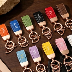 May include: A collection of eleven leather keychains in orange, blue, green, black, red, brown, light blue, purple, yellow, pink, and white. Each features a rose gold metal clasp, a circular ring, and a personalized monogram embossed in gold.
