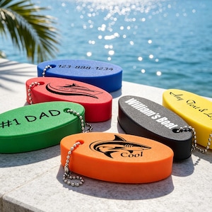 May include: Six oval-shaped foam floating keychains in blue, red, green, orange, black, and yellow. Each keychain features a metal ball chain and custom black text or graphics, including 123-888-1234, Boat life, #1 DAD, a shark, William's Boat, and Any Text & Logo.