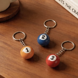 May include: Three billiard ball keychains. The keychains feature a number 1 ball in orange, a number 2 ball in blue, and a number 3 ball in red. Each ball is attached to a silver chain and keyring.